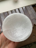 Hand holding polished round Selenite bowl with crystals in background — natural crystal bowl for Crown Chakra energy