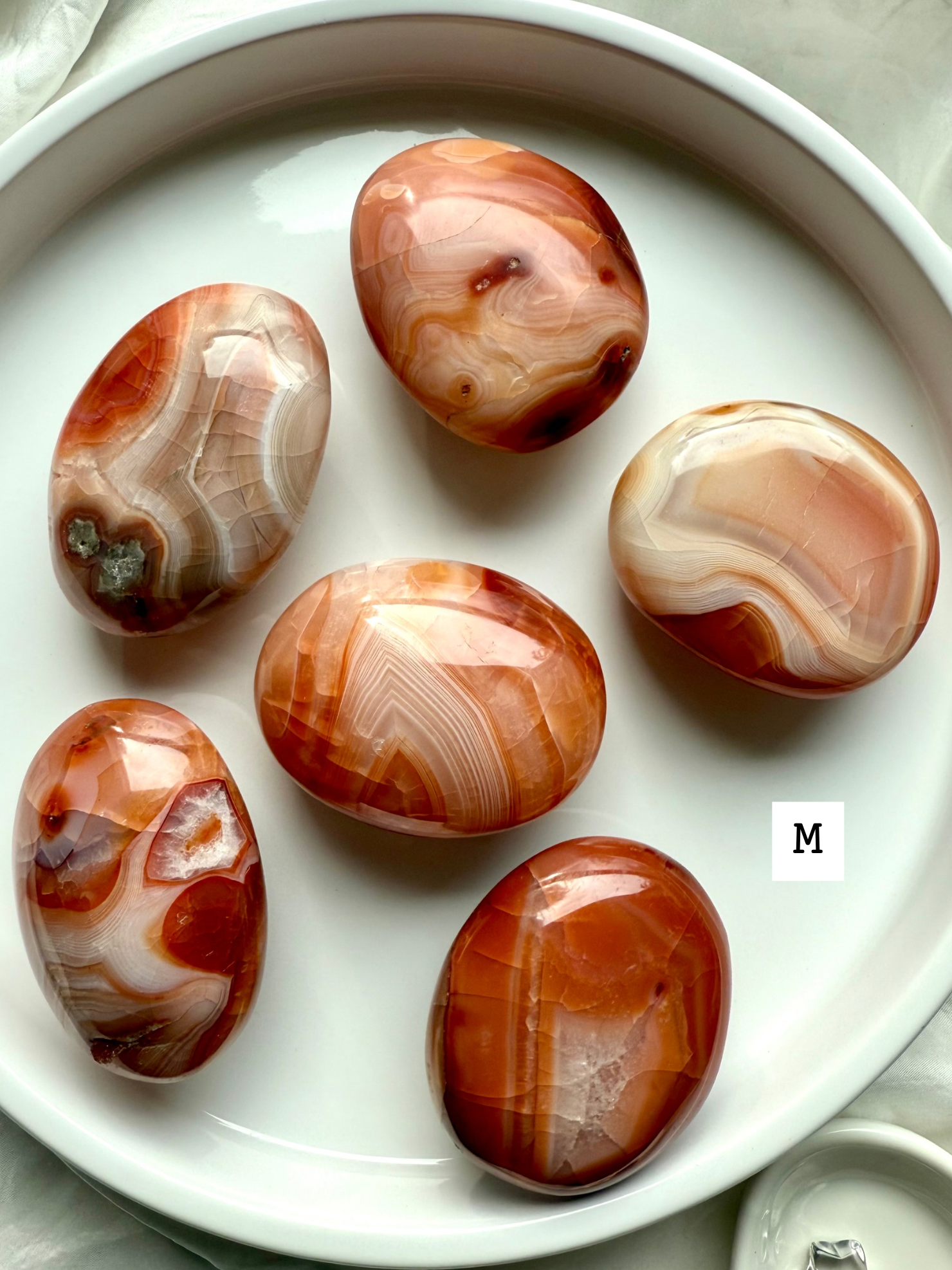 Medium Carnelian palm stones with rich red-orange banding on a white plate — grounding crystals for creativity, confidence + life force energy.
