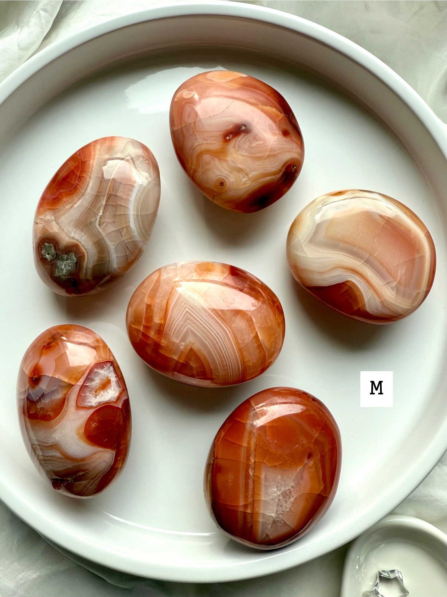 Medium Carnelian palm stones with rich red-orange banding on a white plate — grounding crystals for creativity, confidence + life force energy.