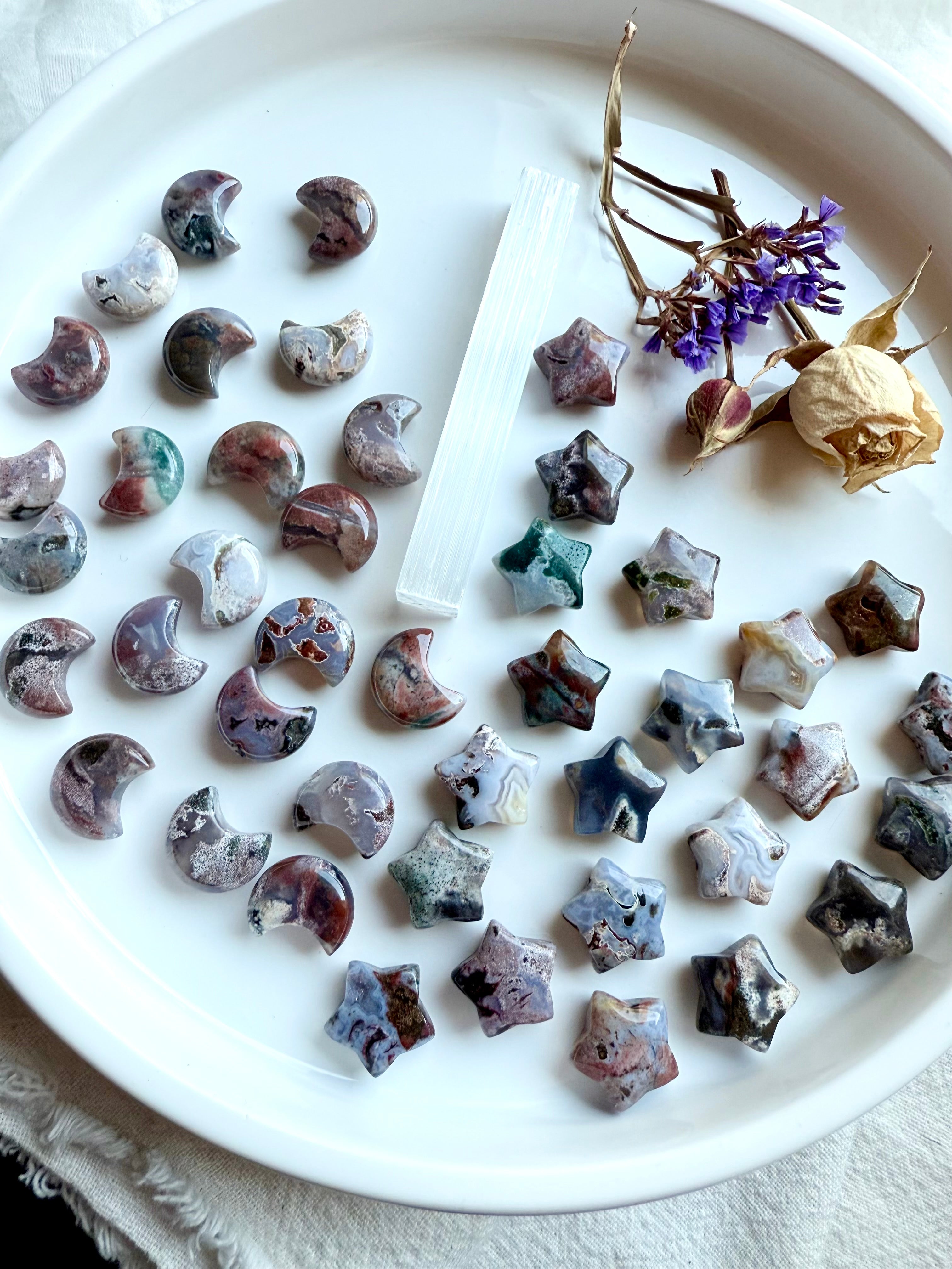 Mini Cosmic Ocean Jasper moons and stars — hand-carved crystal carvings in earthy tones with swirling cosmic patterns on a white dish with flowers and selenite