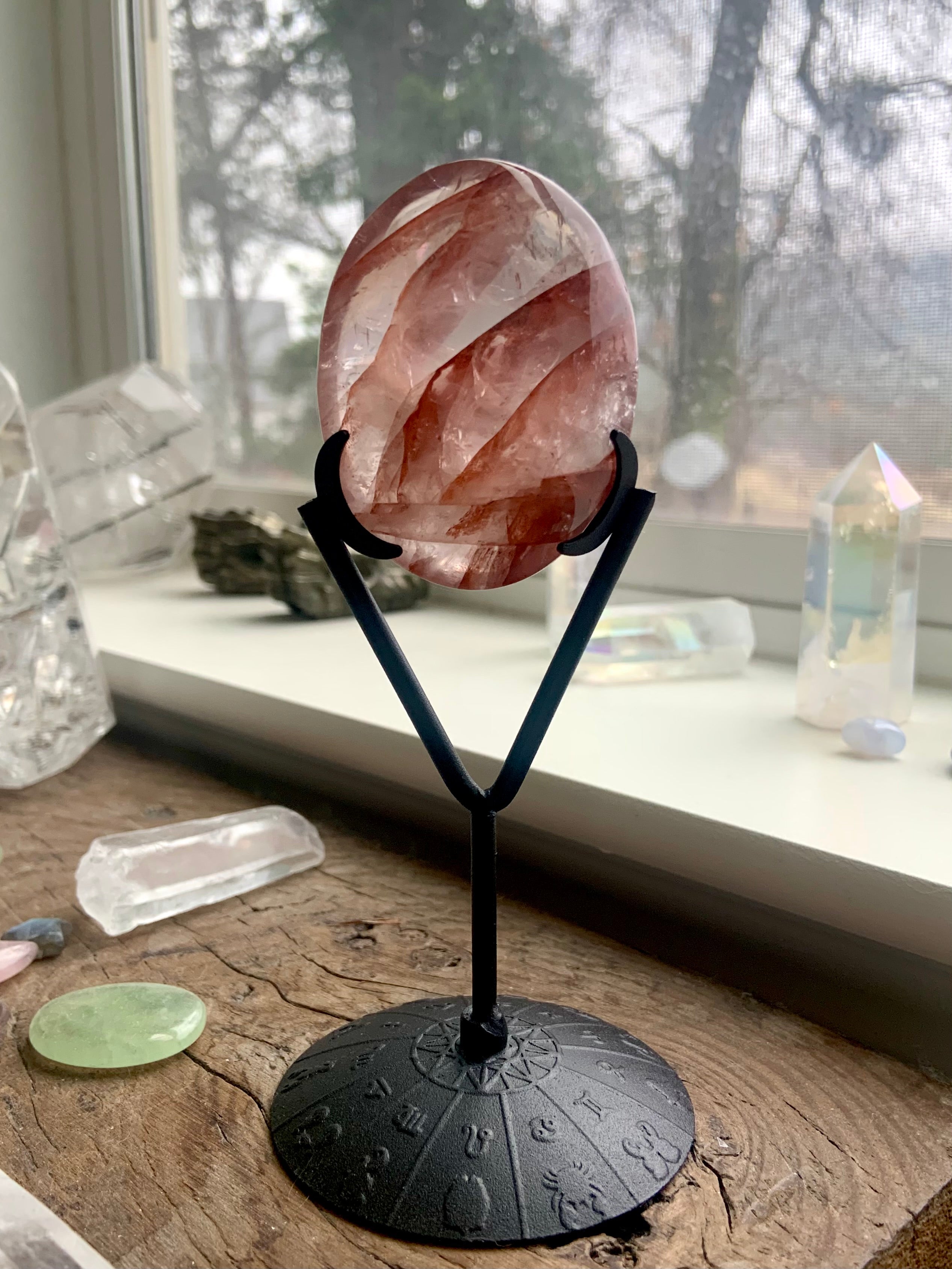 Black crystal display stand holding Fire Quartz palm stone — natural light highlights red hematite ribbons, with cozy windowsill ambiance.