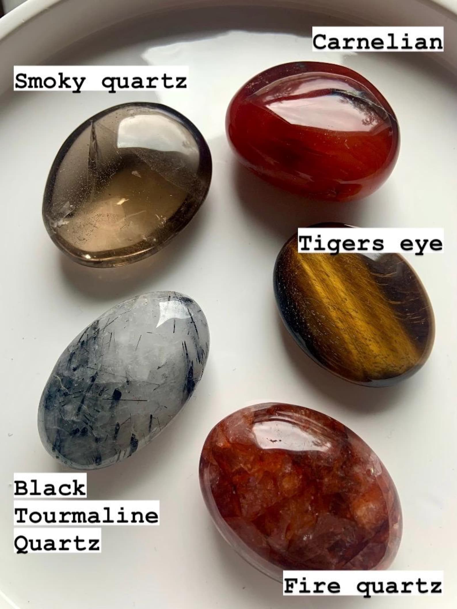 Labeled chakra palm stones on white plate — high-quality polished crystals for lower chakra healing: Smokey Quartz, Tigers Eye, Carnelian, Fire Quartz &amp; Tourmaline in Quartz.