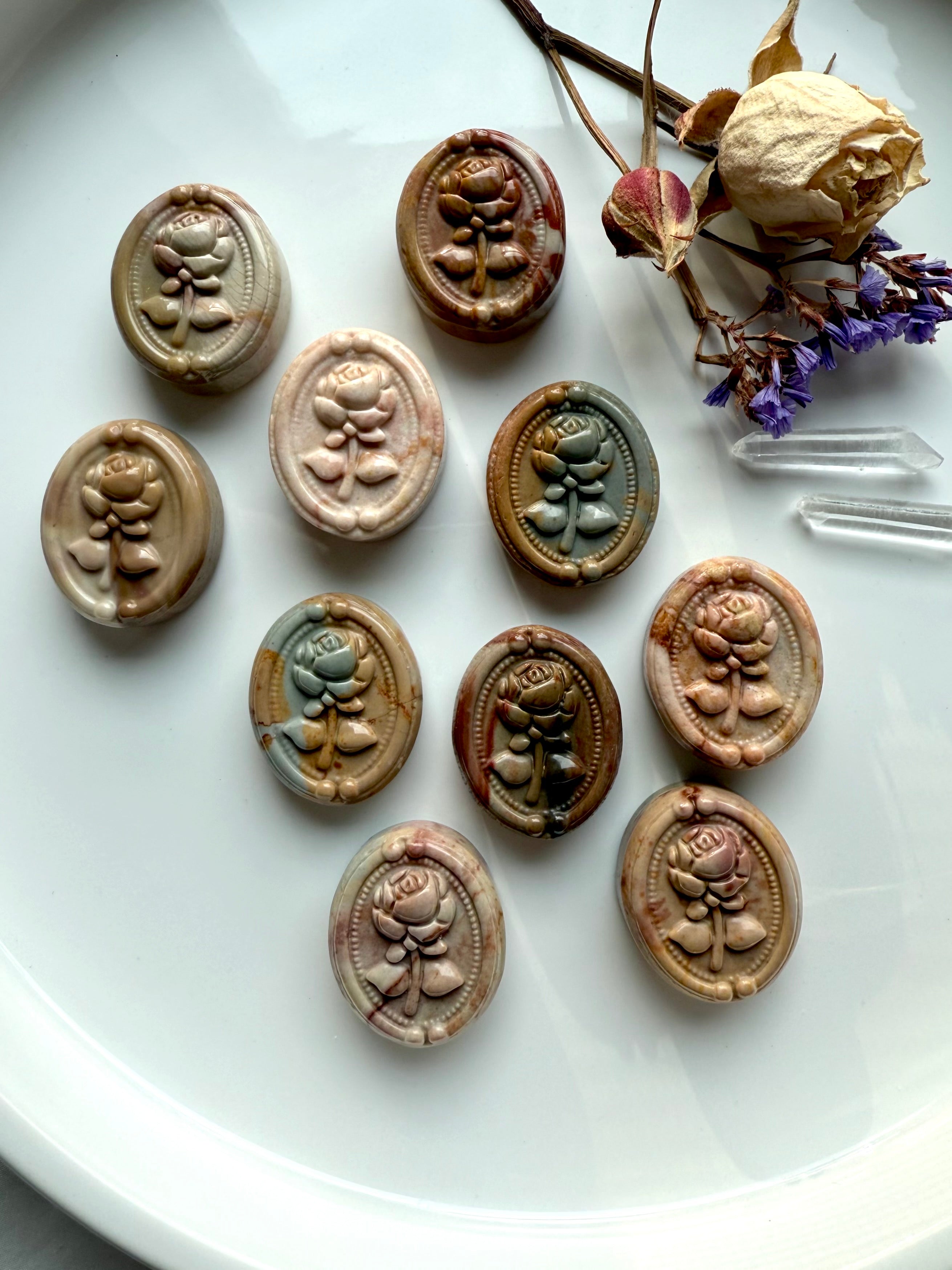 Polychrome Jasper rose cabochons displayed in a group of ten on a white dish with dried flowers and clear quartz points — grounding earth-energy crystals for balance and stability.