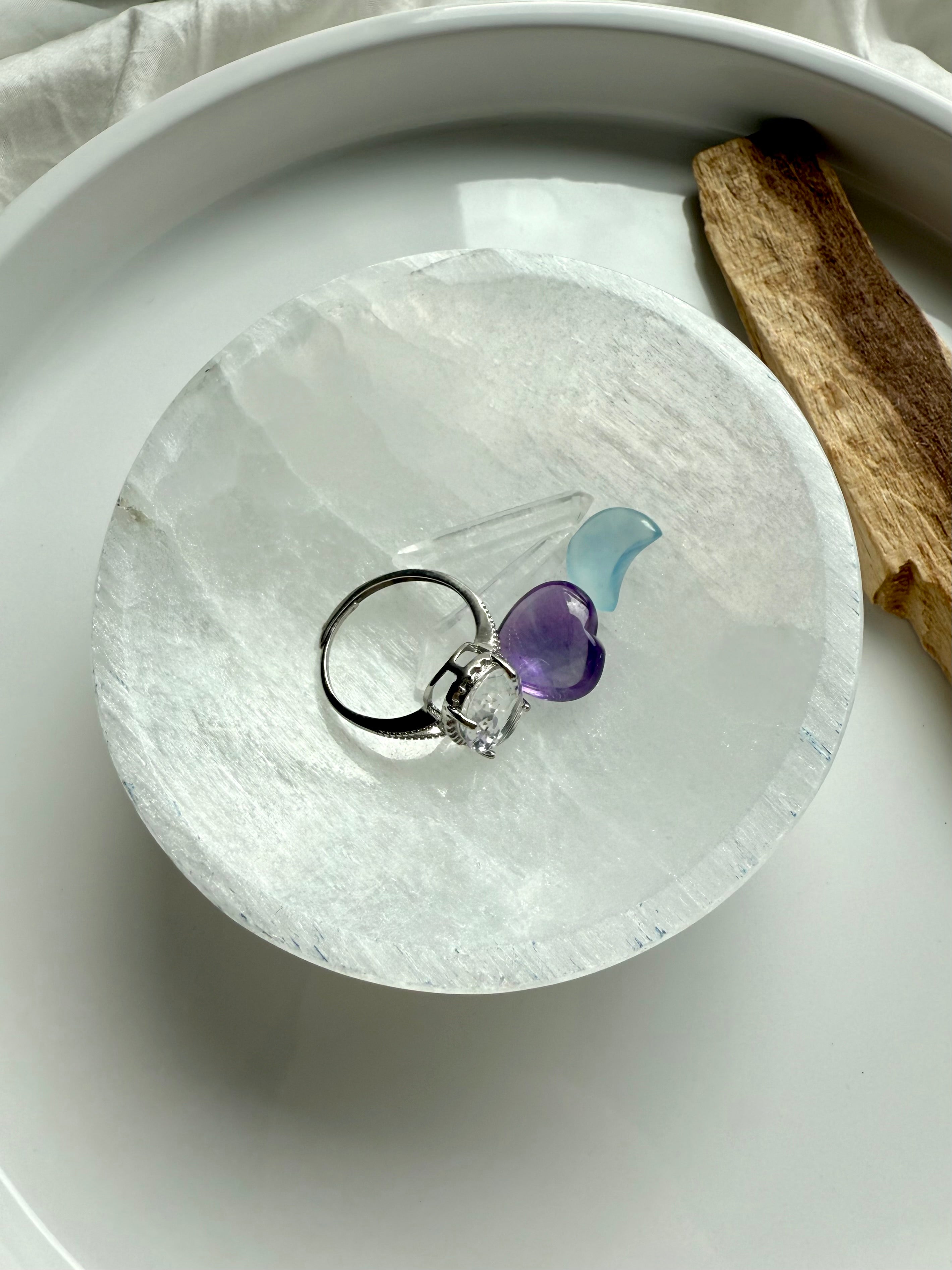 Selenite crystal charging bowl holding a silver ring, amethyst heart, blue crystal, and clear quartz wand on white tray
