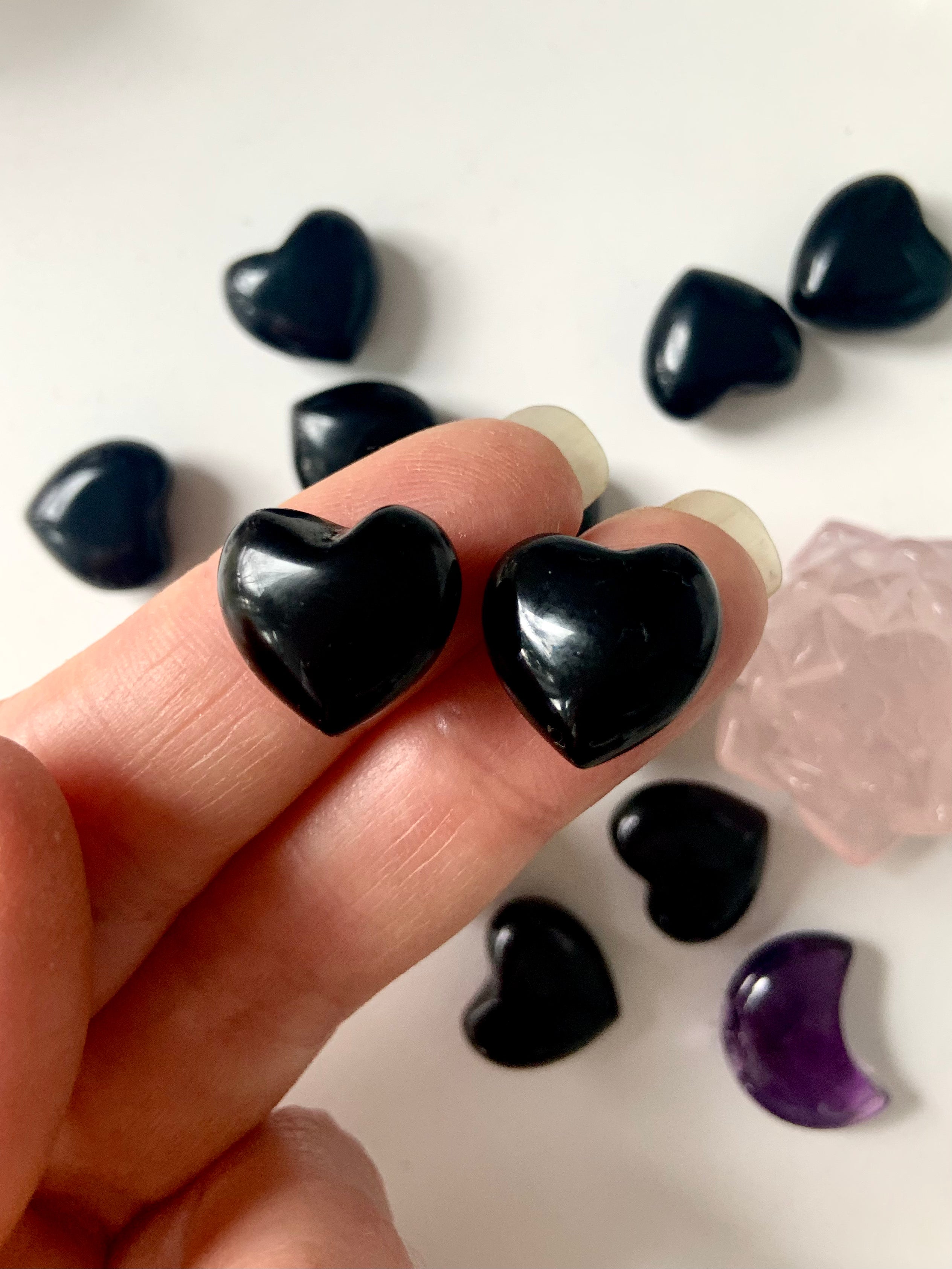 Close-up of two Black Obsidian mini heart carvings held on fingertips with pink and purple crystal accents — powerful pocket talismans