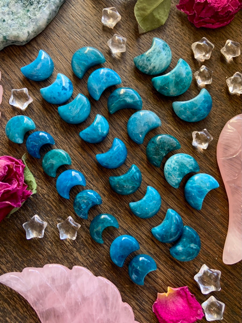 Array of Blue Apatite Crescent Moons arranged by size on wood table — mini moon crystals with rose petals, quartz stars & other carved crystals.