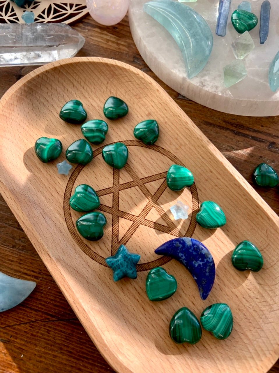 Malachite hearts arranged on a wooden tray with a pentacle engraving, alongside celestial carvings of blue and aqua crystals.