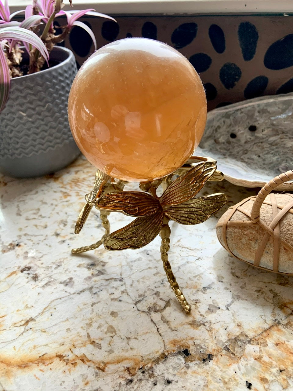 Side view of gold dragonfly crystal stand holding a honey calcite sphere on a marble surface.