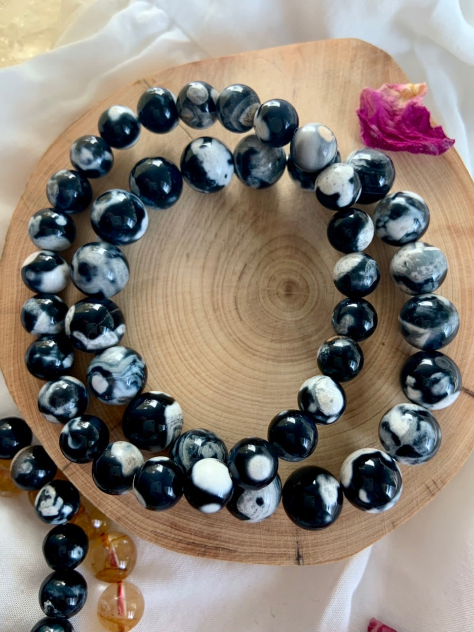 Overhead view of Orca Agate bracelets in 10–11mm and 8–9mm bead sizes resting on a natural wood slice with a pink rose petal for a soft, giftable vibe