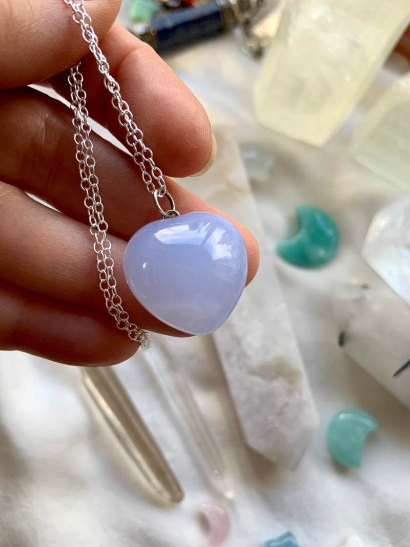 A luminous blue chalcedony puffy heart pendant necklace on s925 silver chain makes a sweet gift her. This pastel blue crystal resonates with the throat + heart chakras. Shown on a hand for scale, with crystals in the background.