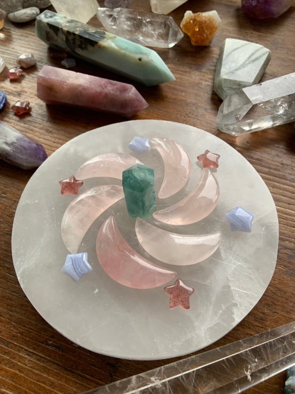 Crystal grid spiral of rose quartz moons, blue lace agate stars, and strawberry quartz stars on selenite plate with other crystals on dark wood table