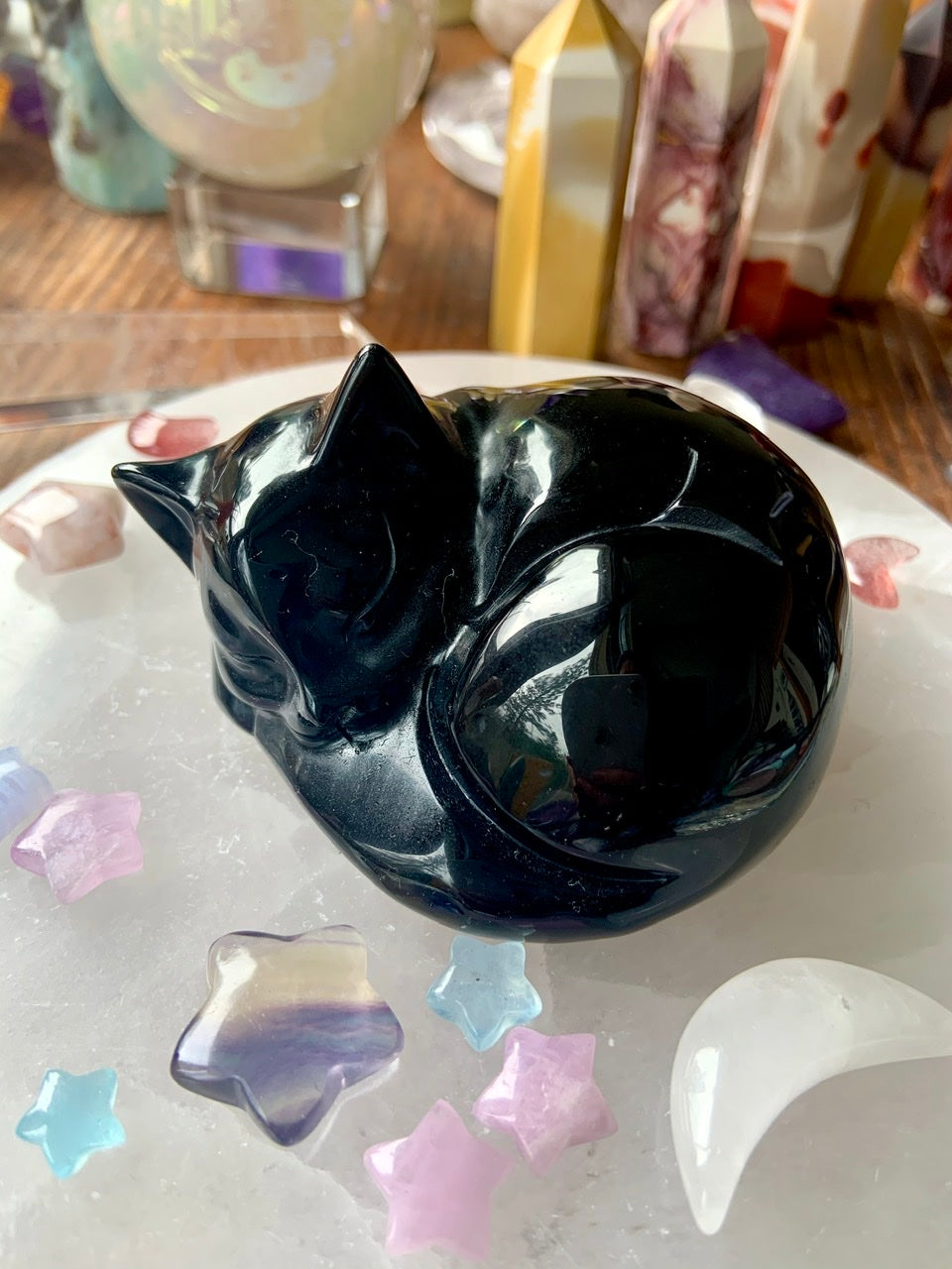Angled view of Black Obsidian sleeping cat figurine resting on a selenite plate, surrounded by pastel celestial crystals — grounding crystal for remembrance.