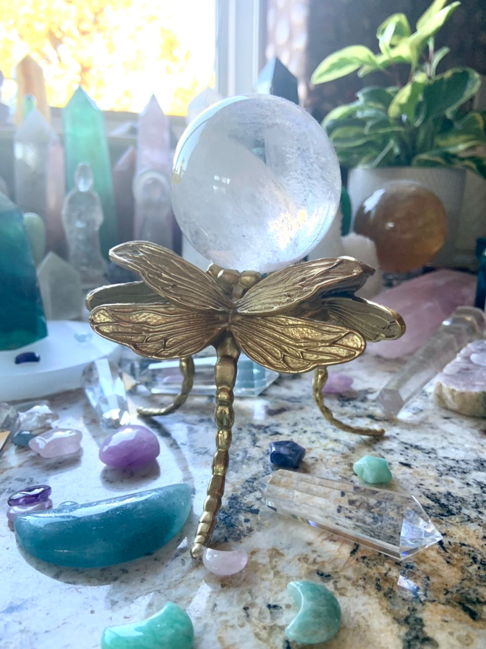 Gold dragonfly crystal ball stand holding a clear quartz crystal sphere, displayed in a colorful crystal collection with plants.