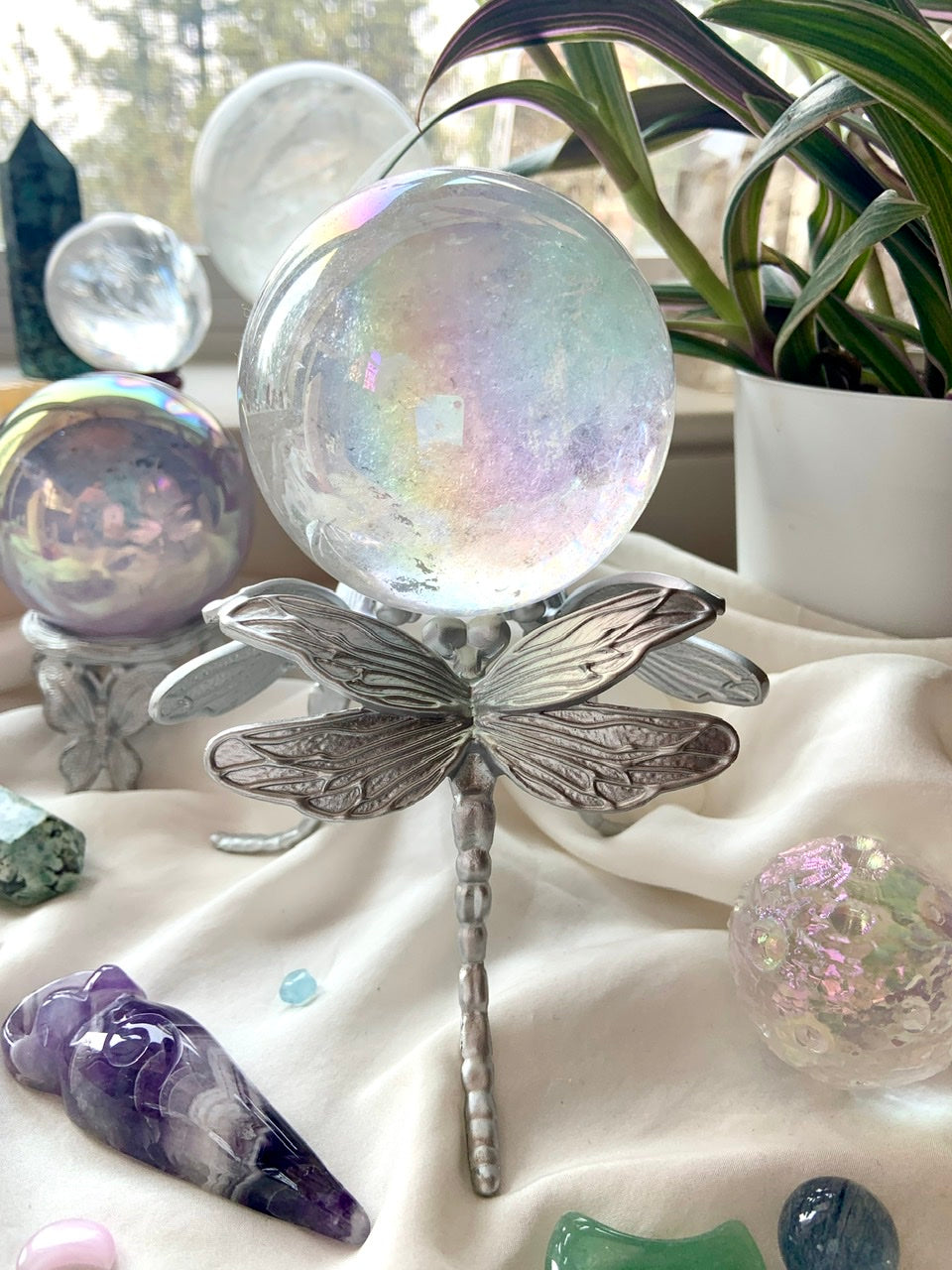 Crystal sphere stand with dragonfly design in silver, holding an aura quartz crystal ball in a dreamy display with plants & backlit crystals.