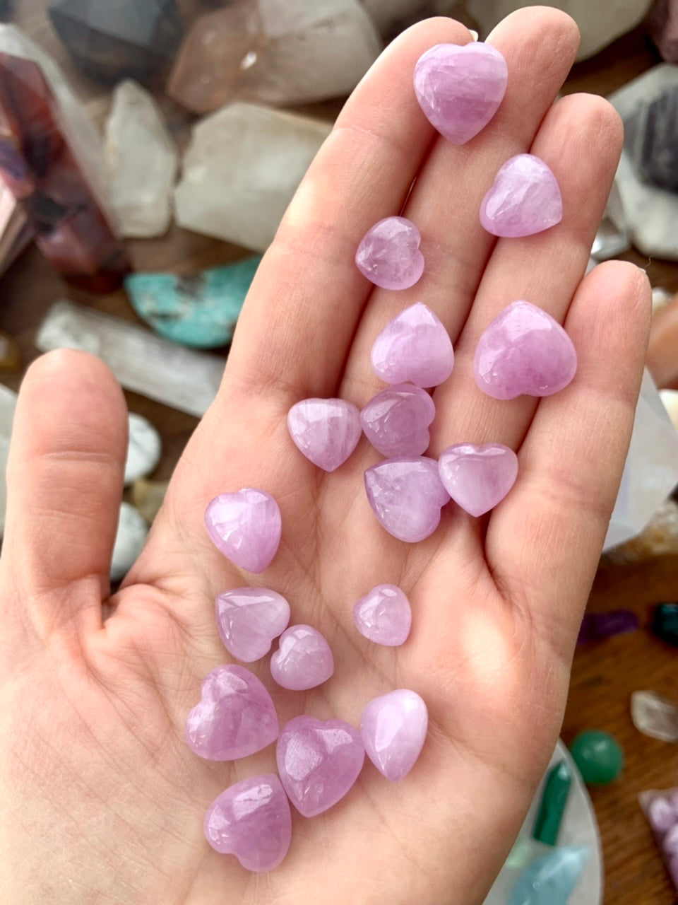 Mini Pink Kunzite hearts displayed in a hand above a crystal-filled table — heart chakra crystals for calm, compassion & soul nourishment.