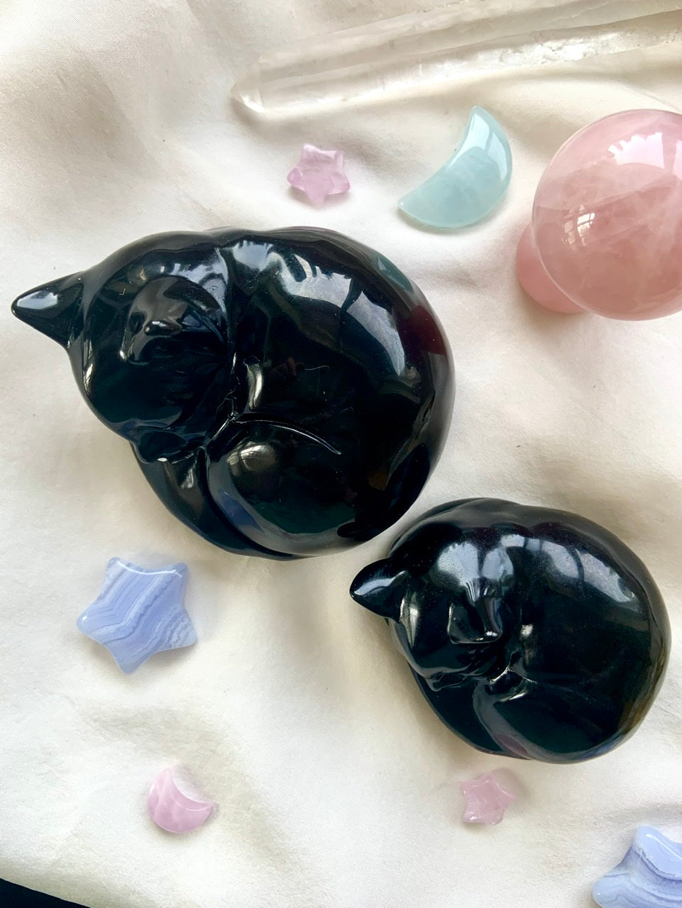 Two Black Obsidian sleeping cat figurines shown with pastel crystals — crystal keepsakes in two sizes for spiritual décor, gifts & pet memorials.