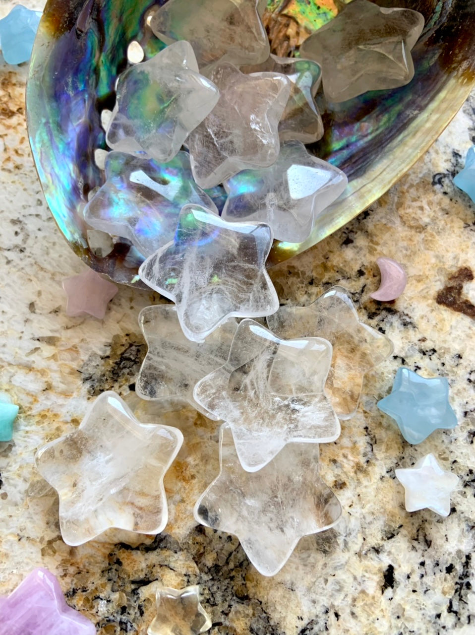 Closeup of Clear Quartz stars spilling out of abalone shell onto granite surface with other celestial crystals, luminous chakra healing carvings