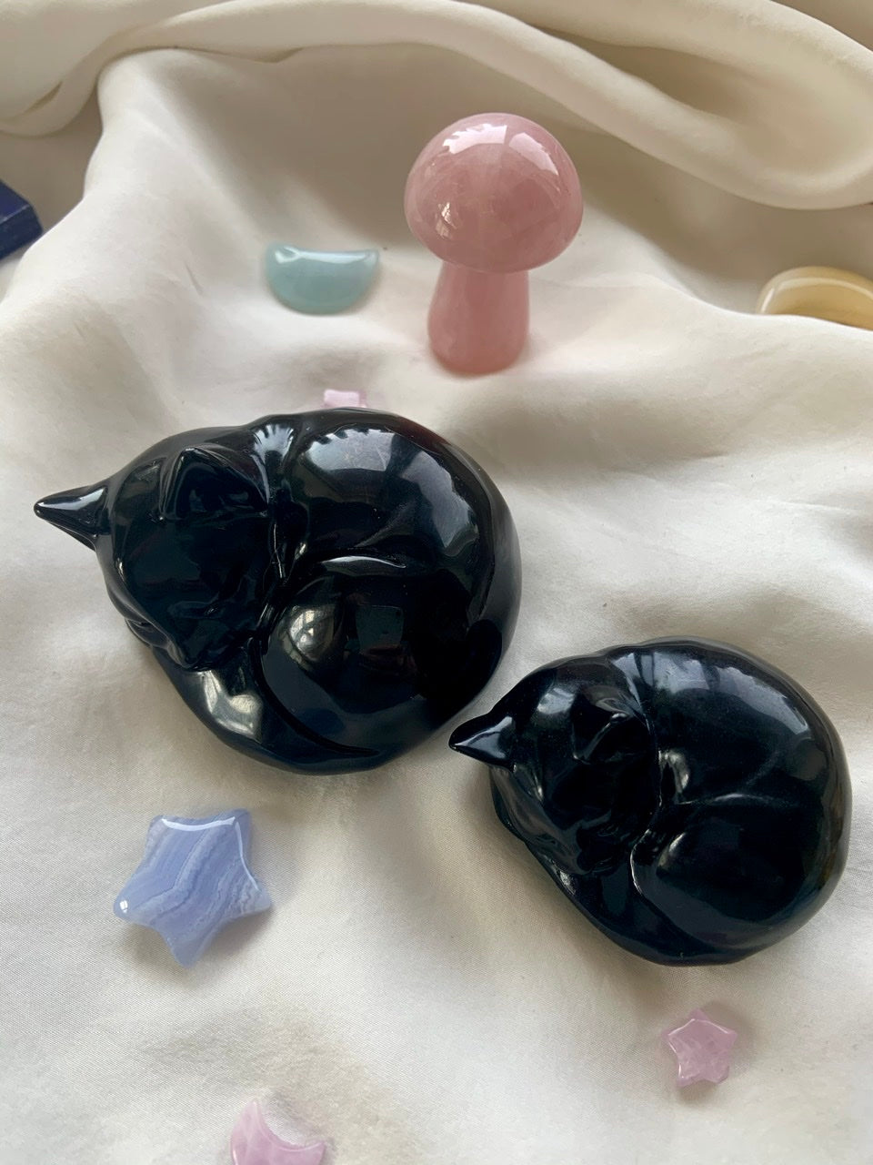 Overhead view of two Black Obsidian Cat carvings on white silk with Rose Quartz mushroom & Blue Lace Agate star — serene crystal arrangement for healing & love.