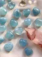 Aquamarine hearts arranged on white silk with a Fire Quartz point — soft blue crystal hearts for emotional healing, clarity + energetic balance.
