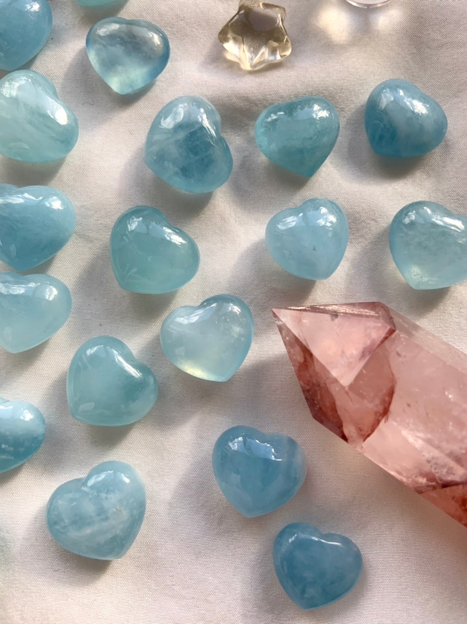 Aquamarine hearts arranged on white silk with a Fire Quartz point — soft blue crystal hearts for emotional healing, clarity + energetic balance.
