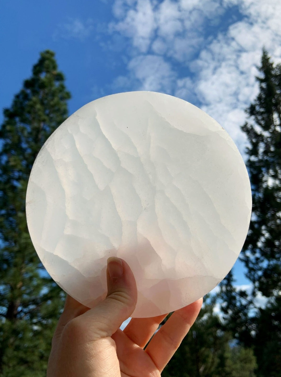 Selenite charging plate held to the light outdoors with blue sky, white clouds, and tree silhouettes
