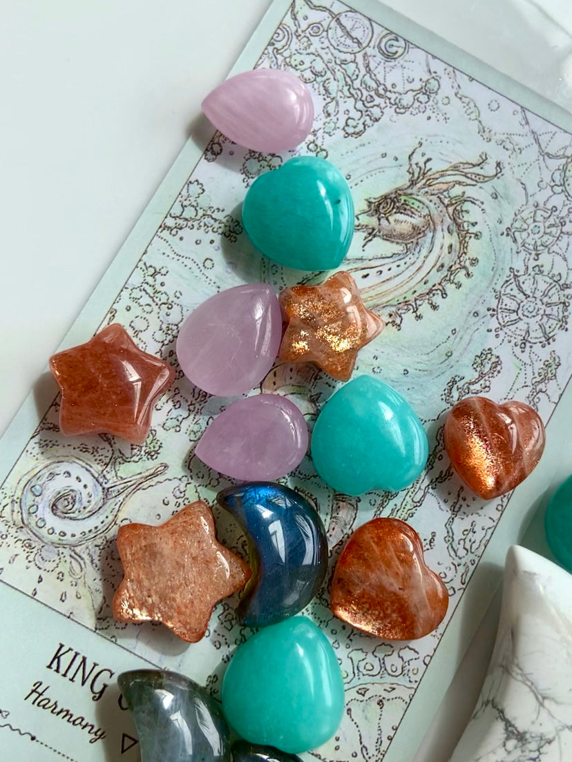 Mini crystal carvings including Sunstone stars, Amazonite hearts, Labradorite moons, & Kunzite teardrops arranged on a tarot card — crystals for healing, intention & energy work.
